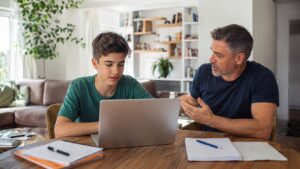 Padre e figlio adolescente conversano davanti a un computer portatile mentre studiano insieme in un soggiorno luminoso.
