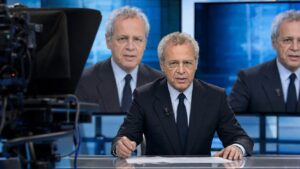 Anchorman dai capelli grigi, somigliante a Enrico Mentana, in uno studio televisivo con il volto alterato digitalmente sui monitor, immagine simbolica dei deepfake satirici.