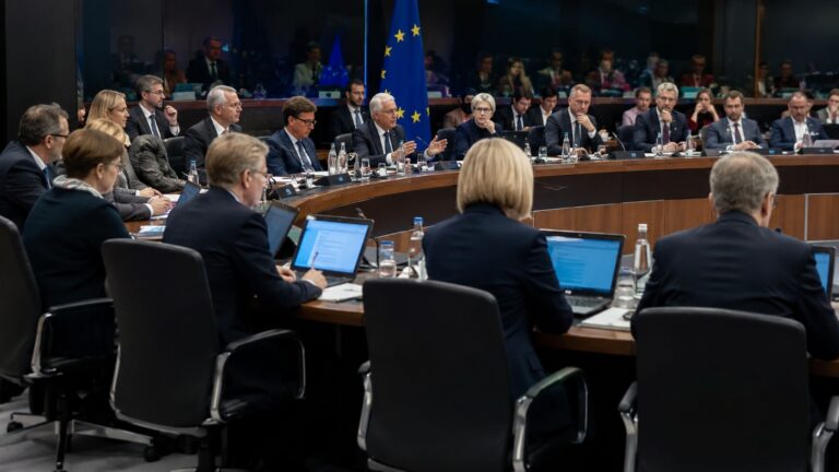 Riunione istituzionale europea con rappresentanti seduti attorno a un grande tavolo, computer e documenti aperti e bandiera dell’Unione europea sullo sfondo, immagine simbolica del confronto regolatorio sull’intelligenza artificiale e Google